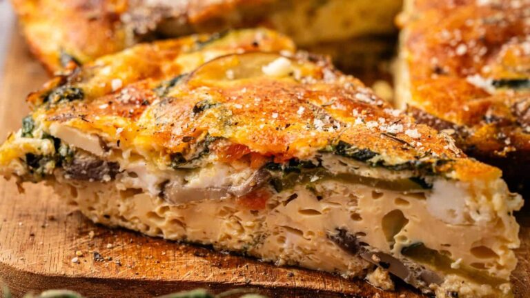 A close-up of a slice of vegetable frittata on a wooden surface, showing layers of egg, vegetables, and seasoning.
