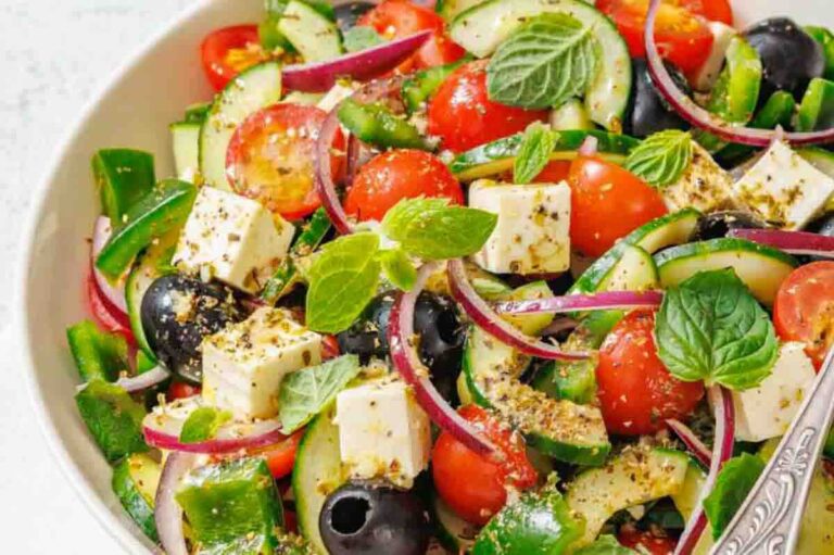 A bowl of Greek salad with chopped cucumber, cherry tomatoes, red onion, green pepper, black olives, feta cheese, fresh mint, and oregano.