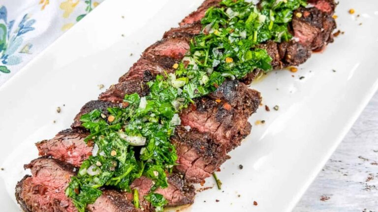 A sliced, medium-rare steak topped with a green herb sauce, served on a rectangular white plate, perfect for any backyard BBQ menu.